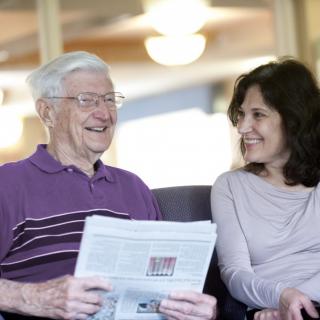 An elderly man and a woman are sitting indoors, smiling and conversing while he holds a newspaper.