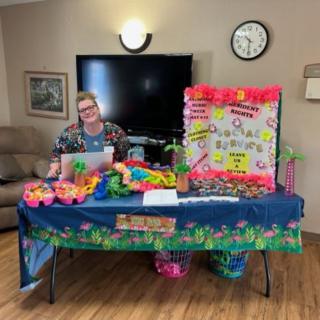 A person sits behind a decorated table with colorful items and a poster indoors.