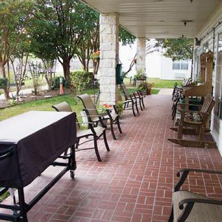 A covered porch with rows of chairs and a brick floor, offering a peaceful outdoor seating area surrounded by greenery.