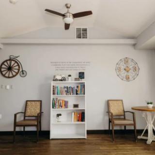 A minimalist living room featuring a white bookshelf and matching chairs with a ceiling fan overhead.