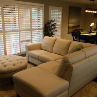 A cozy living room with a cream-colored sectional sofa and round ottoman, accented by natural light filtering through the window shutters.