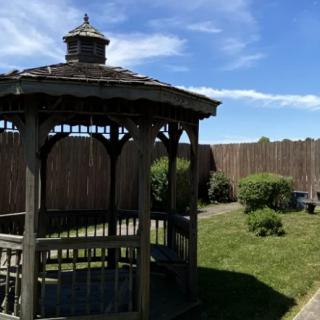 A wooden gazebo in a landscaped garden with surrounding bushes and a wooden fence under a blue sky.