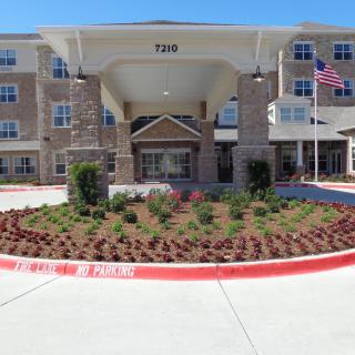 The image depicts the entrance of a multi-story residential building with landscaped gardens and an American flag.