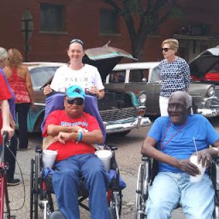 A lively outdoor community event showcasing classic cars, with attendees enjoying the day in wheelchairs.