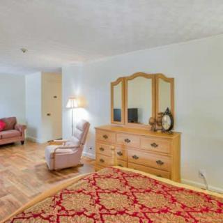 A cozy bedroom with a dresser and mirror, an armchair, a lamp, and a patterned bedspread on a wooden floor.