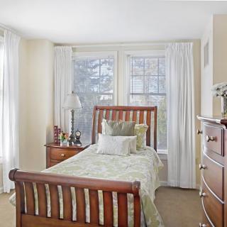 A cozy bedroom with a wooden single bed, nightstand, and dresser, well-lit by natural light from large windows.