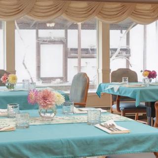 A cozy dining room with tables set in blue tablecloths, adorned with flowers under soft natural light.