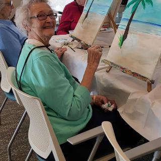 An elderly woman smiling while painting a beach scene on a canvas during an art class.