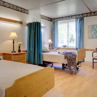 A shared hospital or nursing home room with two beds, a window, and a wheelchair.