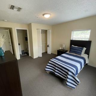 A cozy bedroom with a single bed featuring striped bedding, a nightstand, and a dresser on a carpeted floor.