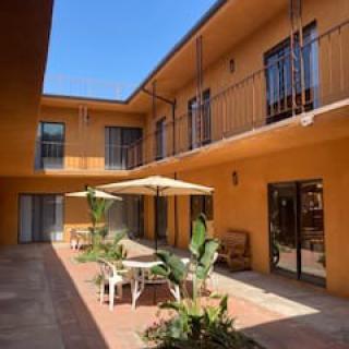 A sunny courtyard with outdoor seating and umbrellas, surrounded by a two-story building with balconies.