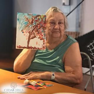 An elderly woman proudly showcasing her painting of a tree, celebrating creativity and artistic expression.
