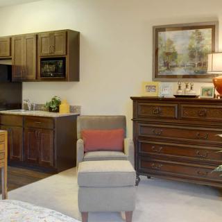 A cozy living area with a kitchenette, featuring a wooden dresser, a comfortable chair, and decorative artwork.
