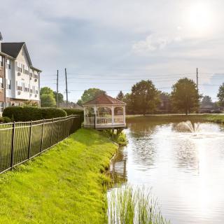A tranquil lakeside view featuring a gazebo, residential building, and lush greenery under a bright sun.