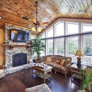 A cozy living room with rustic decor featuring a stone fireplace, wooden ceiling, and large windows.