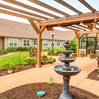 A tranquil garden area featuring a tiered fountain under a wooden pergola, surrounded by well-maintained landscaping and outdoor seating.