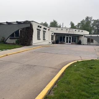 The image shows a building entrance with a paved driveway and surrounding greenery on an overcast day.