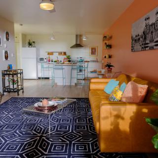 A modern open-plan living room and kitchen with a warm, vibrant color scheme featuring an orange couch, decorative cushions, a geometric-patterned rug, and stylish wall art.