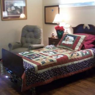 A cozy bedroom featuring a bed with a colorful quilt, an armchair, and a lamp on the bedside table.