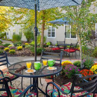 A cozy outdoor patio with tables and chairs under umbrellas, surrounded by lush garden landscaping.
