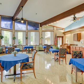 A spacious and airy dining area featuring blue tablecloths, natural light streaming in through large windows, and a welcoming atmosphere.