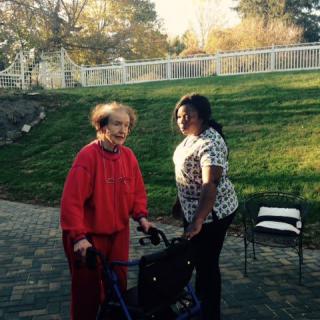 A caregiver assisting an elderly woman with a walker outside on a sunny afternoon.