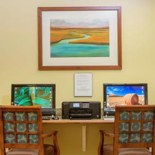 A computer workstation setup with two monitors, a printer, and chairs, featuring artwork on the wall.