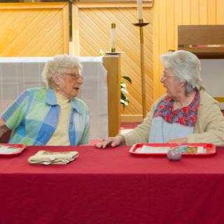Two elderly women engaged in a lively conversation over a meal at a community gathering.