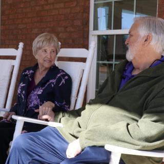 An elderly couple sitting on rocking chairs outdoors, engaged in conversation.