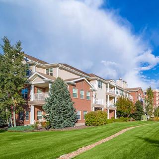 A scenic view of a residential apartment building surrounded by lush grass and trees under a partly cloudy sky.