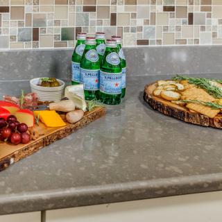 A kitchen counter features a charcuterie board with assorted cheeses, crackers, and grapes, alongside bottles of sparkling water.