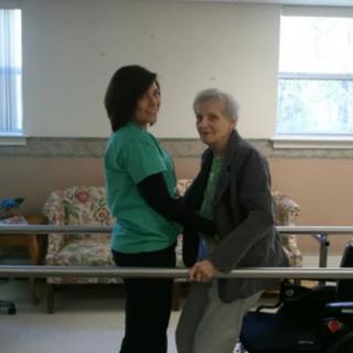 A caregiver assists an elderly woman in a rehabilitation session using parallel bars.
