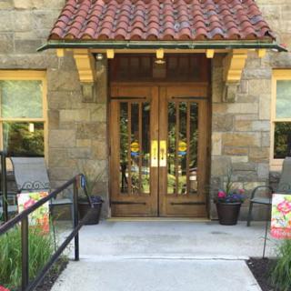 A stone building entrance with wooden doors and a tiled roof, surrounded by plants.