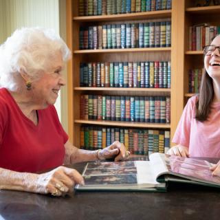 An elderly woman and a young woman share a moment looking through a photo album in a library.