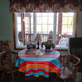 A cozy indoor setting featuring a round table with a colorful tablecloth and surrounded by wooden chairs, placed in front of a large window with a view. The table holds books, a small lamp, and a potted plant.