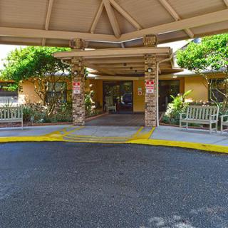 The image shows the entrance of a building with a covered area, benches, and surrounding landscaping.
