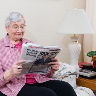 An elderly woman sits comfortably in her living room, reading a newspaper.