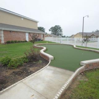 A landscaped pathway with greenery beside a building, fenced area in the background.