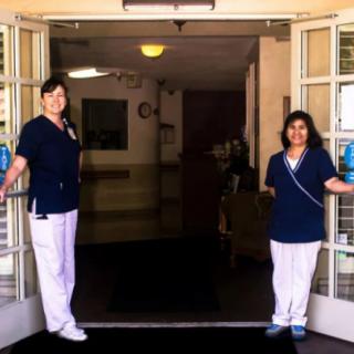 Two nurses stand at the entrance of a healthcare facility, smiling and welcoming visitors.