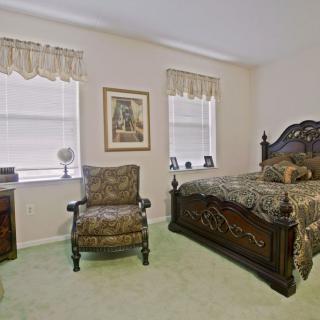 A traditional bedroom features classic furniture with an ornate bed and matching armchair, complemented by natural light from two windows.