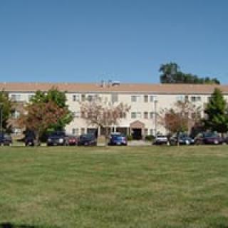 A residential apartment building with a grassy lawn, trees, and parked cars under a clear blue sky.