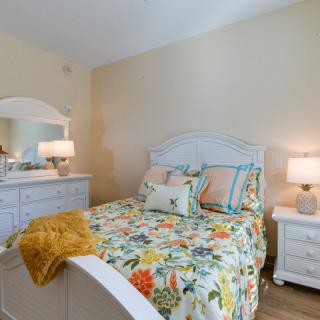 A cozy bedroom with a floral bedspread, featuring a white dresser and mirror, complemented by soft lighting from bedside lamps.