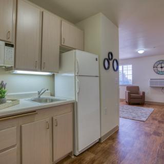 A cozy apartment with a small kitchen featuring white cabinets and a refrigerator, opening into a living area with hardwood flooring.