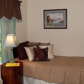 A cozy bedroom with a neatly made bed, soft pillows, and a nearby lamp, featuring elegant curtains and wall art.
