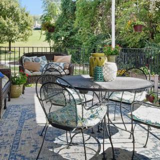 A cozy outdoor patio setup featuring metal chairs and a table, complemented by comfortable cushions and a sofa, surrounded by greenery and decorative plants.