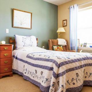 A cozy bedroom with a single bed and a quilt, featuring a nightstand, armchair, lamp, and window.