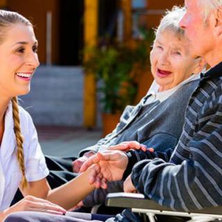 A caregiver interacts compassionately with two seniors seated in wheelchairs outdoors.