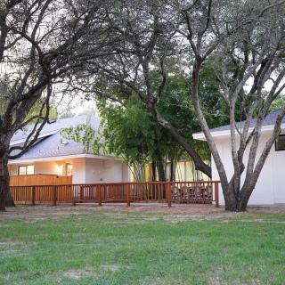 A tranquil backyard featuring a white house with a wooden deck surrounded by lush trees and grass.