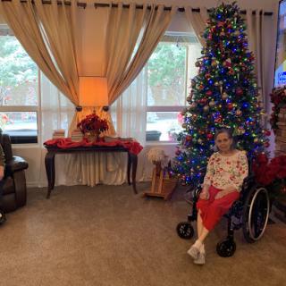 A cozy living room decorated for the holidays, featuring a colorful Christmas tree and a senior citizen in a wheelchair, enjoying the festive environment.