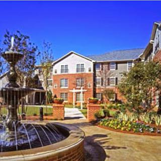 A serene view of a well-maintained apartment complex featuring a beautiful fountain and colorful gardens under a clear blue sky.